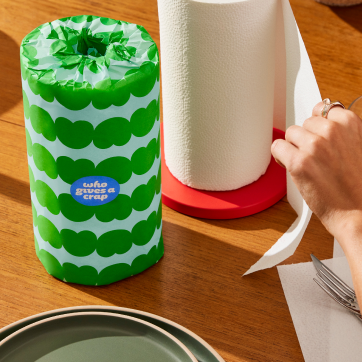 Hand pulling sheet of paper towel, with a colourfully wrapped paper towel sitting ontop of a tabletop.