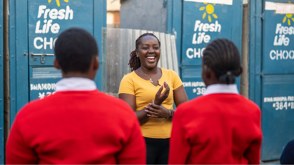 A smiling young woman standing in front of a Fresh Life toilet block with two children in school uniform in front of her.