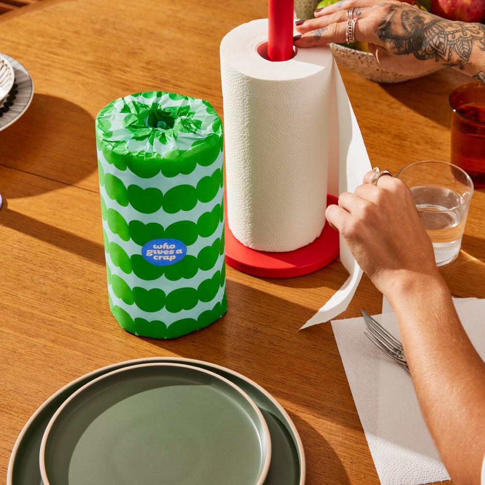 A person is tearing sheets from a roll of kitchen towels next to a green wrapped roll of kitchen towels on a table top.