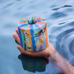 A hand holding a single Who Gives A Crap Splash Edition bamboo toilet roll above a body of water, showing illustrated swimmers and bathers on the colourful wrapper