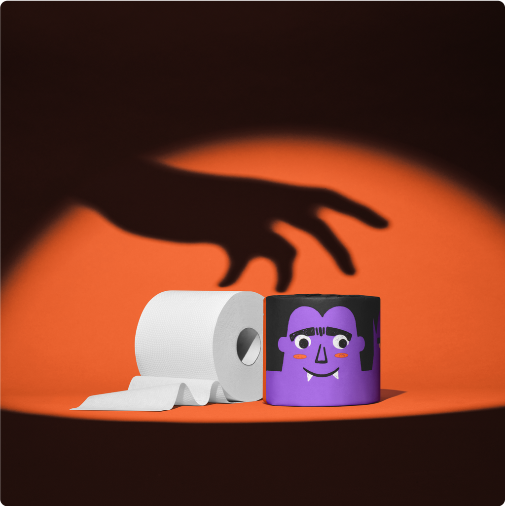 Purple vampire roll of Spooky Edition toilet paper beside an unrolled white 3-ply roll, with a creeping hand shadow