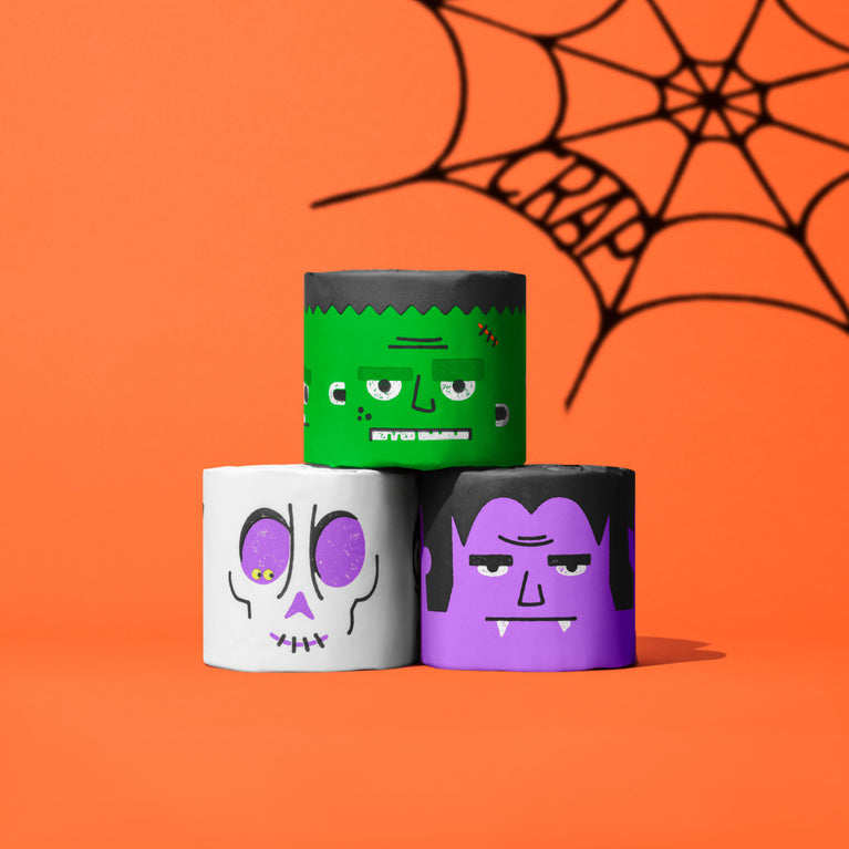 Three Spooky Edition toilet paper rolls stacked in Frankenstein, skull and vampire designs against an orange spider web backdrop