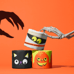Skeleton hand lifting a mummy-print toilet paper roll above black cat and pumpkin rolls on orange
