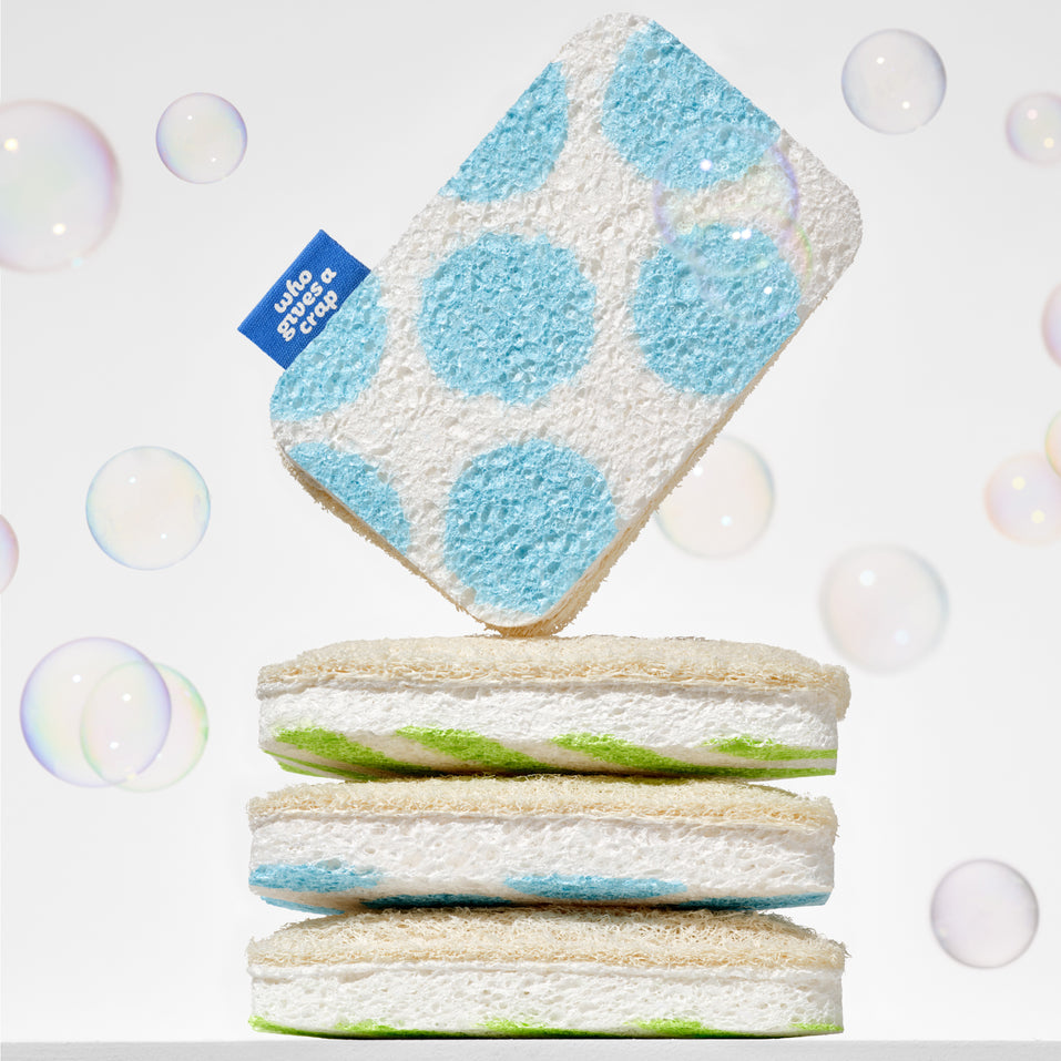 A Who Gives A Crap blue polka dot scourer sponge balanced on top of a stack of green striped sponges surrounded by soap bubbles