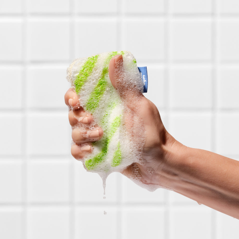 A hand squeezing a lathered Who Gives A Crap green striped scourer sponge against a tiled wall, with soapy water dripping down