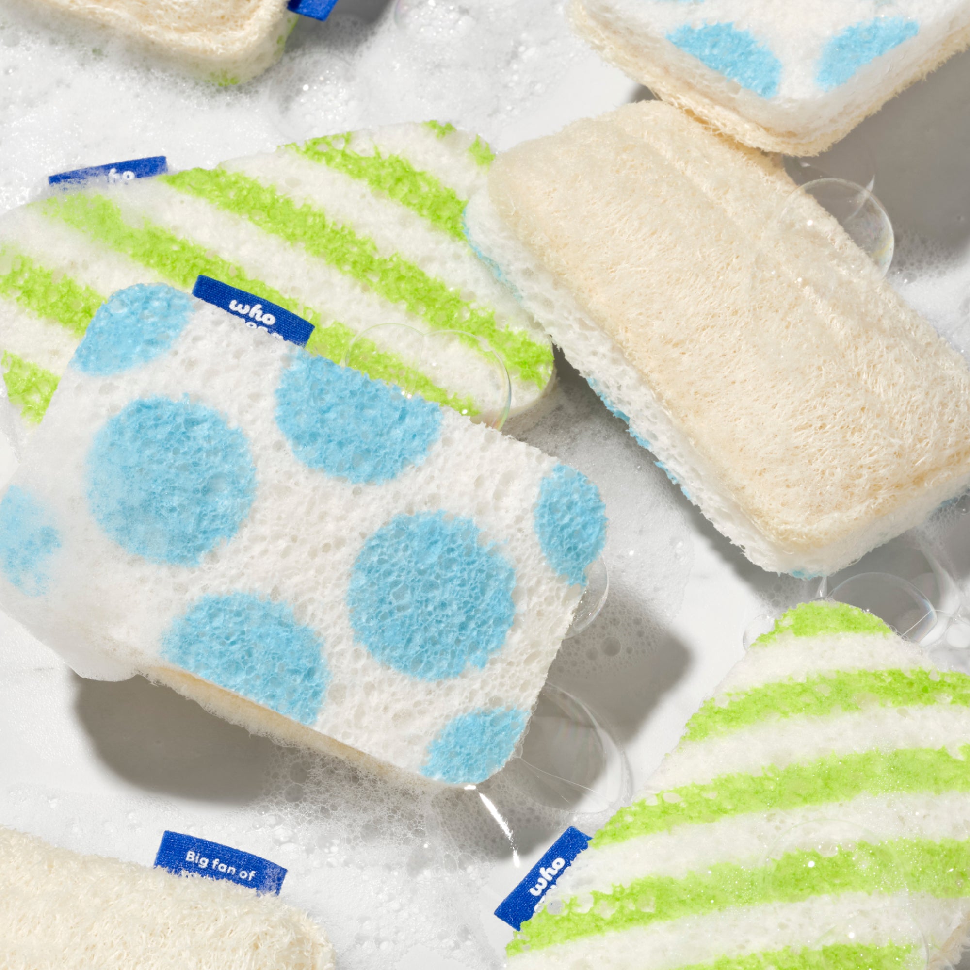 Multiple Who Gives A Crap scrub sponges floating in soapy water, showing the blue polka dot, green stripe and natural loofah designs