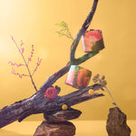 Two nature-themed wrapped toilet paper rolls are balanced on twisted branches against a warm yellow background. One roll has an image of a pink cockatoo and the other has an image of a red banksia