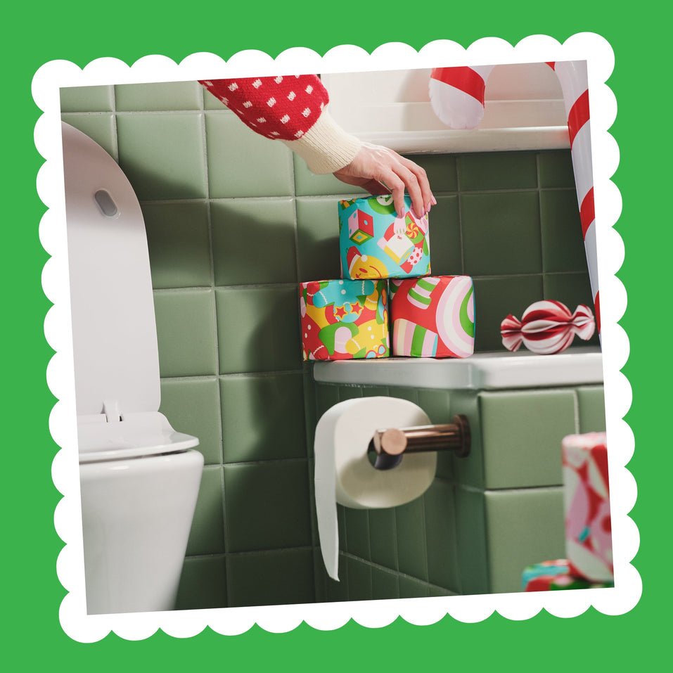 A hand places a wrapped Gift Edition roll on a stack of festive rolls on a bathroom shelf above a toilet roll holder.