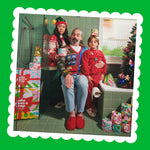 A family in festive jumpers sits on a toilet in a green-tiled bathroom surrounded by Gift Edition boxes, wrapped rolls and a Christmas tree.
