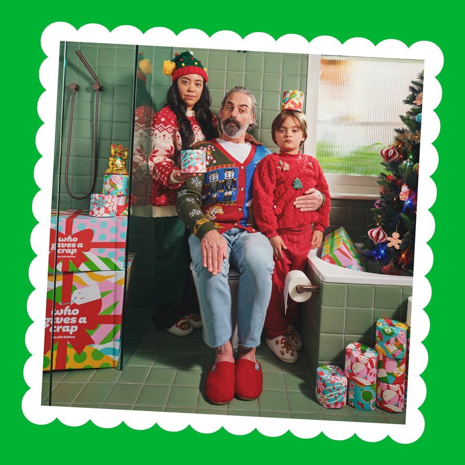 A family in festive jumpers sits on a toilet in a green-tiled bathroom surrounded by Gift Edition boxes, wrapped rolls and a Christmas tree.