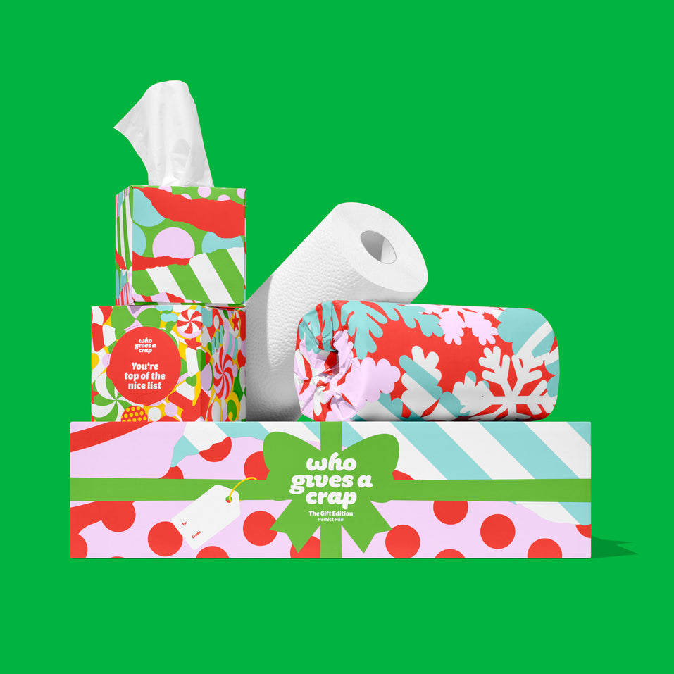 The Perfect Pair: Who Gives A Crap Gift Edition tissue boxes and wrapped paper towel rolls in festive red, pink, green and blue designs, stacked on a gift box on a green background.