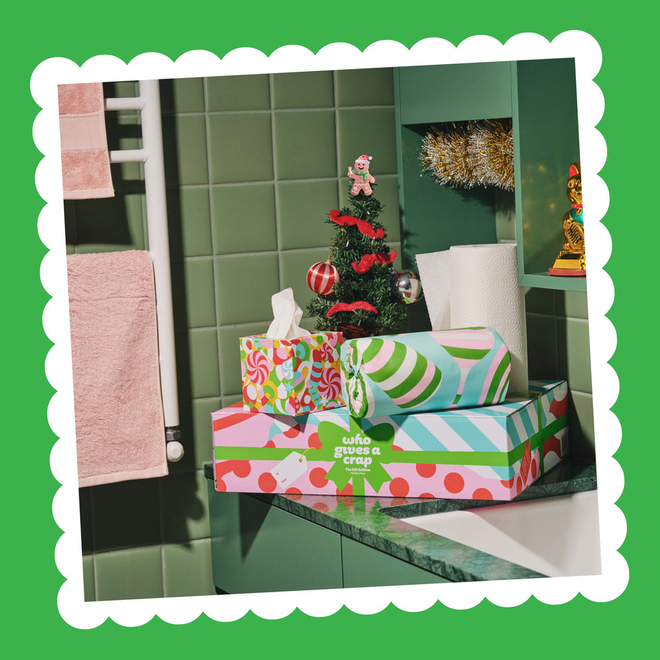 Perfect Pair tissue boxes and paper towel rolls in festive wrappers on a green marble bathroom bench beside a small Christmas tree.
