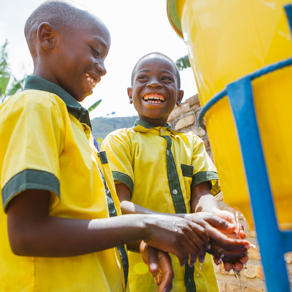 Two boys in yellow school uniforms laughing as they wash their hands together at a water tank in Rwanda