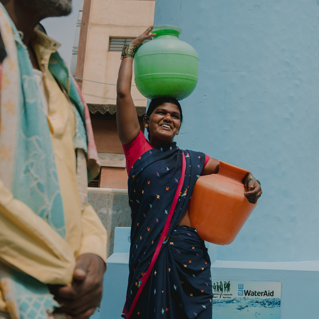 Walking woman carries containers of water on head and hip and smiles