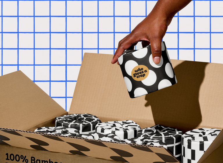 Hand reaches into box of Who Gives a Crap toilet paper holding a single roll of bamboo toilet paper