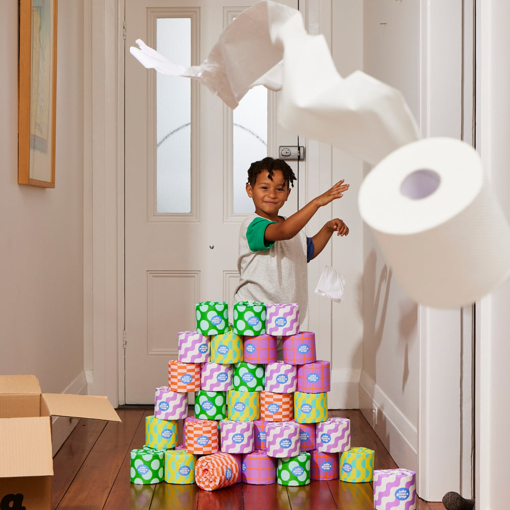 Young smiling boy standing in front of colourful stack of Who Gives A Crap recycled toilet paper playfully throwing toilet paper roll at camera.